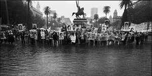 Historische schwarz-weiß-Aufnahme von Menschen, die mit Plakaten mit den Fotos ihrere verschwundenen Angehörigen auf einem überfluteten Platz in Buenos Aires stehen