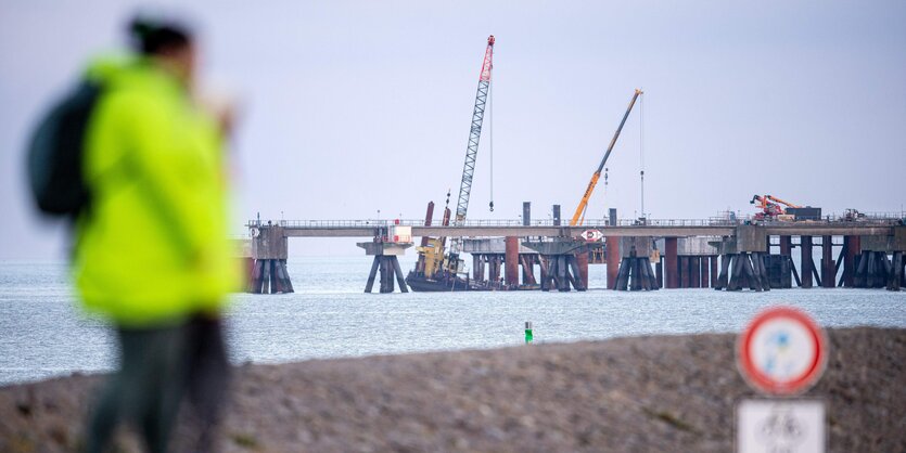 Kranschiffe am LNG-Terminal in Wilhelmshaven. Eine Person in gelber Neonjacke schaut vom Land aus aufs Meer und das Terminal