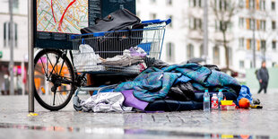 Schlafsack und andere Habseligkeiten eines Obdachlosen rund um einen Einkaufswagen neben einem öffentlichen Stadtplan am Bremer Hauptbahnhof