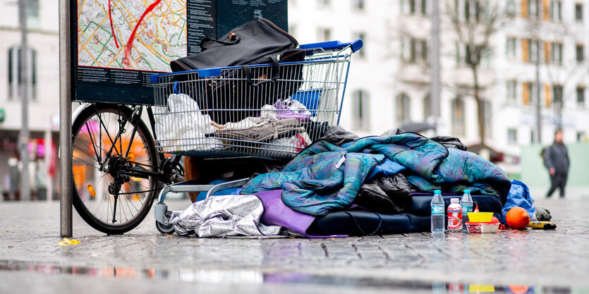 Schlafsack und andere Habseligkeiten eines Obdachlosen rund um einen Einkaufswagen neben einem öffentlichen Stadtplan am Bremer Hauptbahnhof