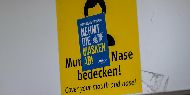 Die Aufforderung, eine Maske zu tragen, ist mit einem Aufkleber: Nehmt die Masken ab, verdeckt