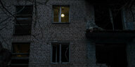 Eine beschädigte Hausfassade eines Mehrfamilienhauses, in einem Fenster leuchtet eine Deckenlampe.