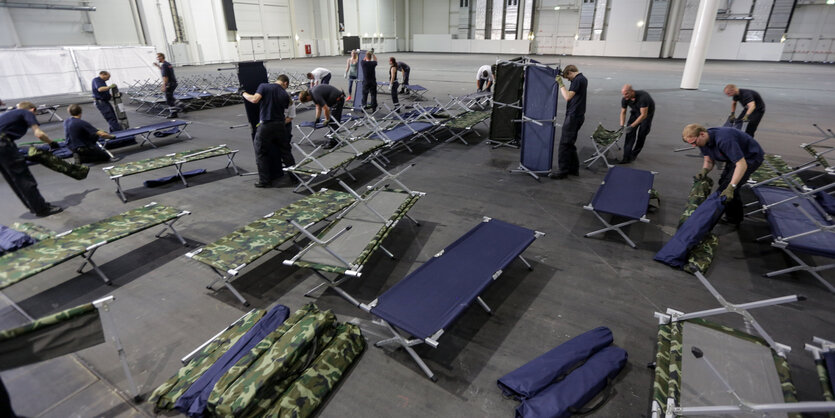 Helfer stellen in einer Halle Feldbetten auf