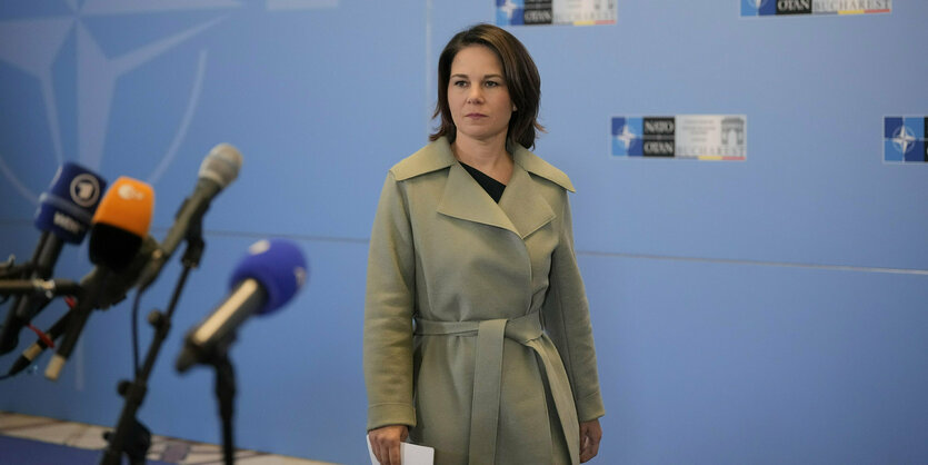 Annalena Baerbock im Trenchcoat mit Blättern in der Hand, beim Betreten eines Raumes für eine Pressekonferenz, im Hintergrund Nato-Farben an der Wand, im Vordergrund ein Pult mit Mikrofonen.