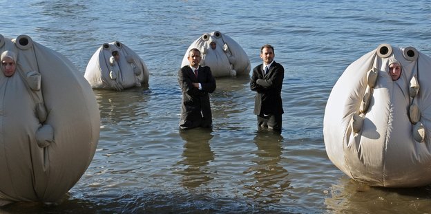 In große runde Säcke gekleidete Menschen in knietiefem Wasser, dazwischen zwei entnervt schauende Anzugträger