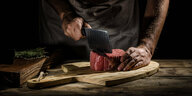 Männerhände mit einem Hackebeil über einem großen Steak auf einem Holzbrett