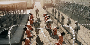 Szene aus dem Film „Fünf Jahre Leben“. Männer in orangenen Häftlingsanzügen und mit schwarzen Säcken über den Köpfen knien auf einem umzäunten Platz.