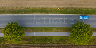 Das Luftbild von einer Drohne zeigt eine Landstraße, auf die jemand Markierungen und das Wort "DB-Trasse" gepinselt hat. Am Straßenrand steht ein rot-weißes Kreuz als Zeichen des Protestes.