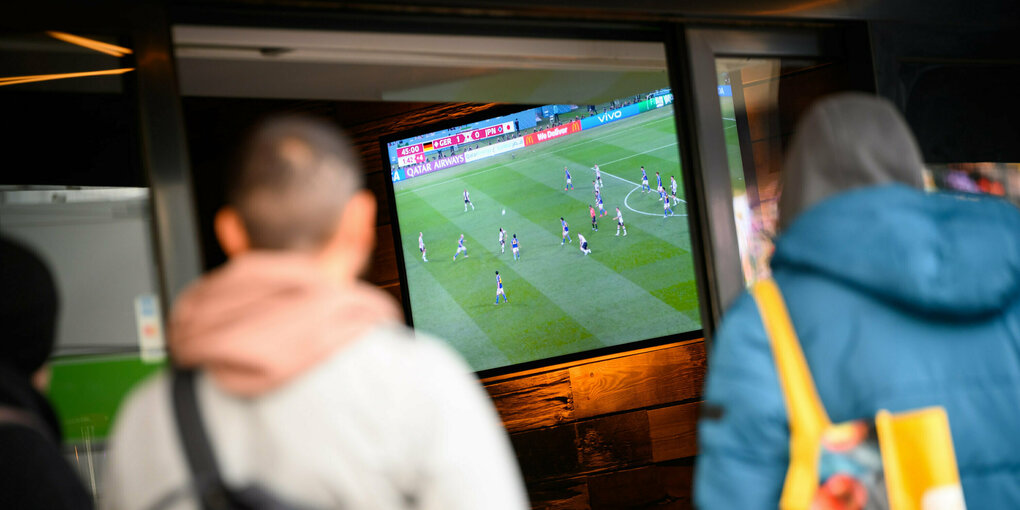 Zwei passanten vor einem Bildschrim, auf dem Fußball läuft