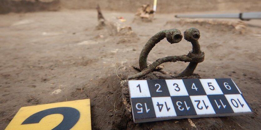 Eine Hufeisenfibel liegt im Sand, davor zwei Schilder