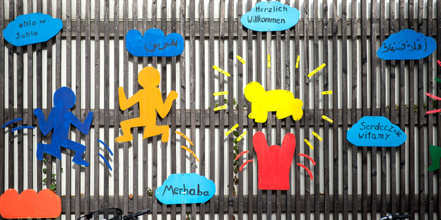 Auf dem Zaun einer Schule sind bunte Figuren und das Wort "Willkommen" in mehreren Sprachen