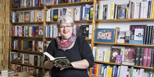 Die Buchhändlerin Sylvia Pyrlik steht in einem schwarzen Kleid vor Regalen und hält ein aufgeschlagenes Buch in der Hand