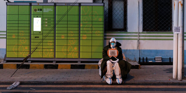 Ein Mitarbeiter der Anti-Corona Kräfte in Peking sitzt mit Schutzkleidung unterm Wintermantel vor einem Haus