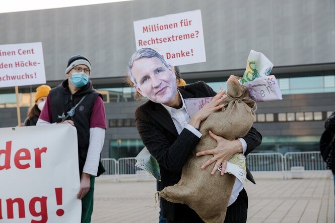 Menschen protestieren mit Schildern, einer hat sich eine Maske aufgesetzt, die das Gesicht von Björn Höcke zeigt