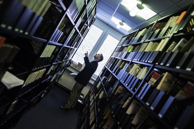 Blick in eine Bibliothek, vor einem Fenster sieht man die Silhouette eines Mannes