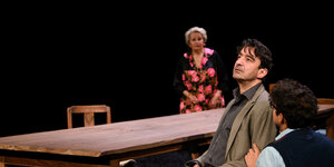 Szene aud dem Bühnenstück "Die Vögel", Hauptfigur Eitan sitzt am Tisch, im Hintergrund seine Mutter Leah