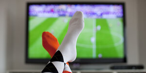 Überschlagene Beine vor einem Fernsehgerät, auf dem ein Fußballspiel übertragen wird.