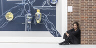Ulrike Kremeier leitet das Brandenburgische Landesmuseum für moderne Kunst, sie sitzt am Boden in einem Museum in Cottbus, hinter ihr an der Wand ein abstraktes Gemälde