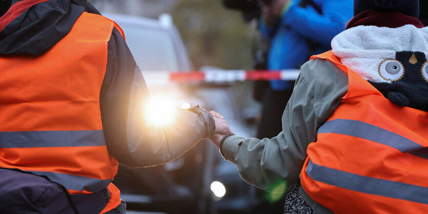 Zwei Personen in Warnwesten sitzen einem Auto gegenüber und halten sich an den Händen