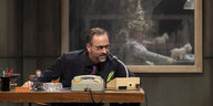 Kurt Krömer sitzt in seiner TV-Show an einem alten Schreibtisch und hinter ihm wartet hinter einer Glasscheibe ein Gast
