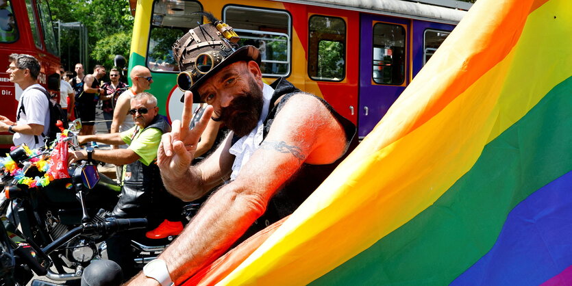 Eine Person hinter einer Regenbogenfahne grüßt in die Kamera