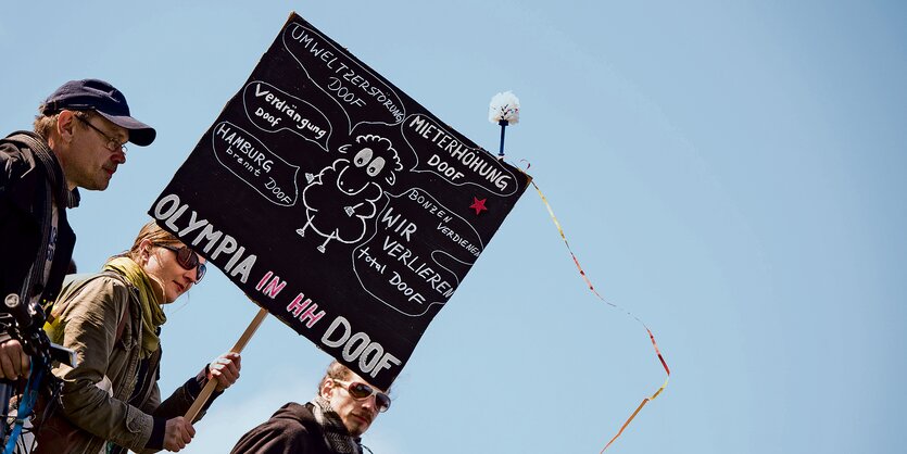 Drei Menschen in dunkler Kleidung vor blauem Himmel, eine Person trägt ein schwarzes Pappschild