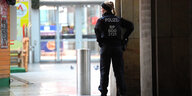 Rückenansicht einer Polizistin mit blondem Zopf, die in einiger Entfernung zum Eingang einer Shopping Mall steht, daneben eine Weihnachtsbude