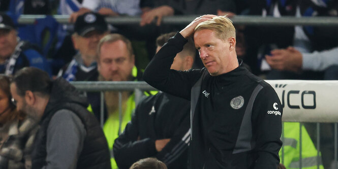 Ein Mann in schwarzer Trainingsjacke steht am Rand des Spielfeldes und fasst sich an den Kopf