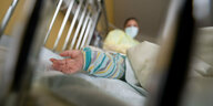 Babyhändchen ragt aus einem Gitterbett, Pflegerin im Hintergrund