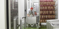 In einer weißen Halle in einer Fleischfabrik schiebt eine Frau einen Ständer mit vielen aufgehangenen Würsten umher