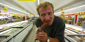 Ein gestelltes Symbolfoto eines Mannes mit Schnauzer, der im Supermarkt in eine Wurst beißt