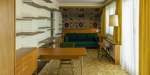 Ein Raum mit hellem Parkettboden, Regalen und Tisch. Das grüne Sofa sthet an der Stirnwand in einer Nische aus hellem Holz und einem kirgiesischen Wandteppich