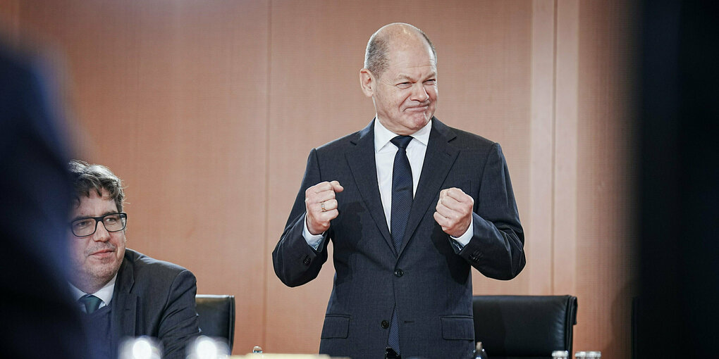 Olaf Scholz hebt die Fäuste und presst mit einer Grimasse die Lippen aneinander, dahinter eine braune Holzwand