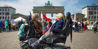 Zwei Menschen im Rollstuhl, dahinter eine Gruppe Demonstrierender und das Brandenburger Tor