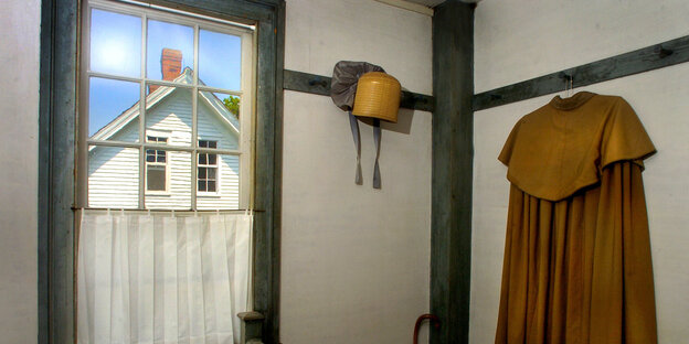 Ein brauner Umhang hängt an einem Haken, durch das Fenster ist der Giebel eines hellen Holzhauses zu sehen