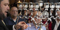 Ein Rabbiner zündet zusammen mit einem weiteren Mann die erste Kerze einer Chanukkia. Im Hintergrund versammeln sich Menschen und gucken den Beiden zu.