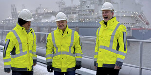 Robert Habeck, Olaf Scholz und Christian Lindner stehen in gelben Warnjacken gekleidet am Ufer, hinter ihnen ein zugeschneites Schiff