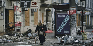 Eine mit Trümmern übersäte Stadtstraße, eine ältere Frau geht im Mantel auf dem Gehsteig, im Hintergrund verrammelte Häuser