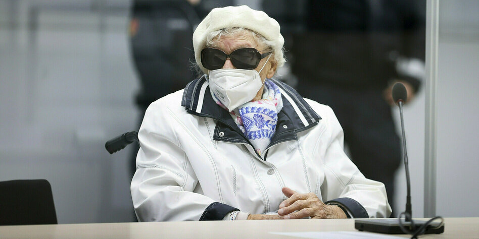 Eine alte Frau auf der Anklagebank eines Gerichtssaals, das Gesicht ist mit einem Mund-Nasenschutz und einer großen Sonnenbrille verdeckt