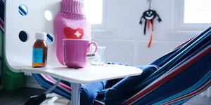 Fieberthermometer, Hustensaft, Wärmflasche und eine Tasse Tee stehen auf einem Stuhl, während ein Kind im Hintergrund in einer Hängematte liegt