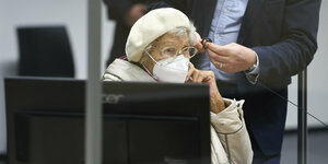 eine alten Frau sitzt hinter einem Bildschirm auf der Gerichtsbank, sie trägt beige Kleidung und eine Mütze und Maske