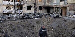 Ein Journalist steht in einem Krater vor einem zerstörten Hochhaus