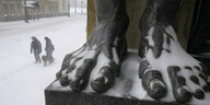 Füße einer verschneiten Statue, im Hintergrund zwei Menschen im Schneesturm