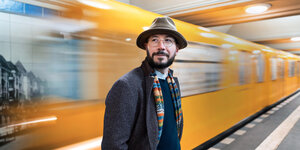Ein Mann mit Hut steht vor der gelben U-Bahn, die vorbeizieht