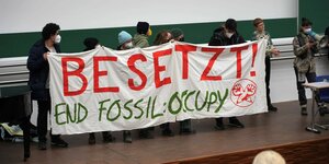 Junge Menschen stehen in einer Reihe vor einem grünen Hintegrund im Audimax in Leipzig. Sie halten einen weißen Banner mit der Aufschrift "Besetzt! End Fossil: Occupy