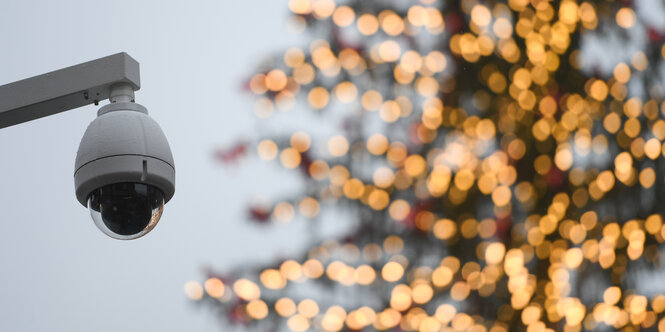 Links im Vordergrund eine Überwachungskamera, die an ein Pfeiler hängt. Im Hintergrund ein Tannenbaum mit gelb leuchtender Lichterkette.