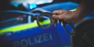 Eine Hand hält Handschellen vor einem Polizeifahrzeug