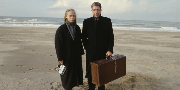 Leonard Cohen mit einem Koffer in der Hand neben einer Frau am Strand