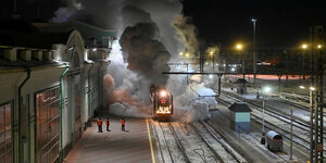 Eine winterlich geschmückte Dampflok fährt in einen Bahnhof ein und macht riesige Rauchwolken