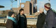 Drei Männer und eine Frau stehen in Winterkleidung auf einem Bahnsteig, gegenüber das VW Werk in Wolfsburg mit dem großen VW Logo an der Fassade